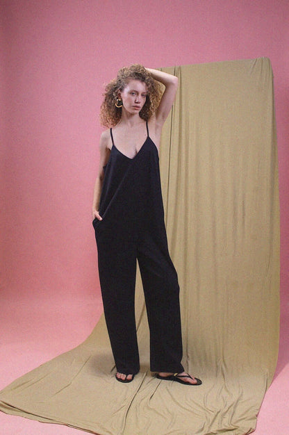 Model wearing Organic Cotton Wide Jumpsuit in Black, featuring slim straps and a relaxed fit against a pink backdrop.