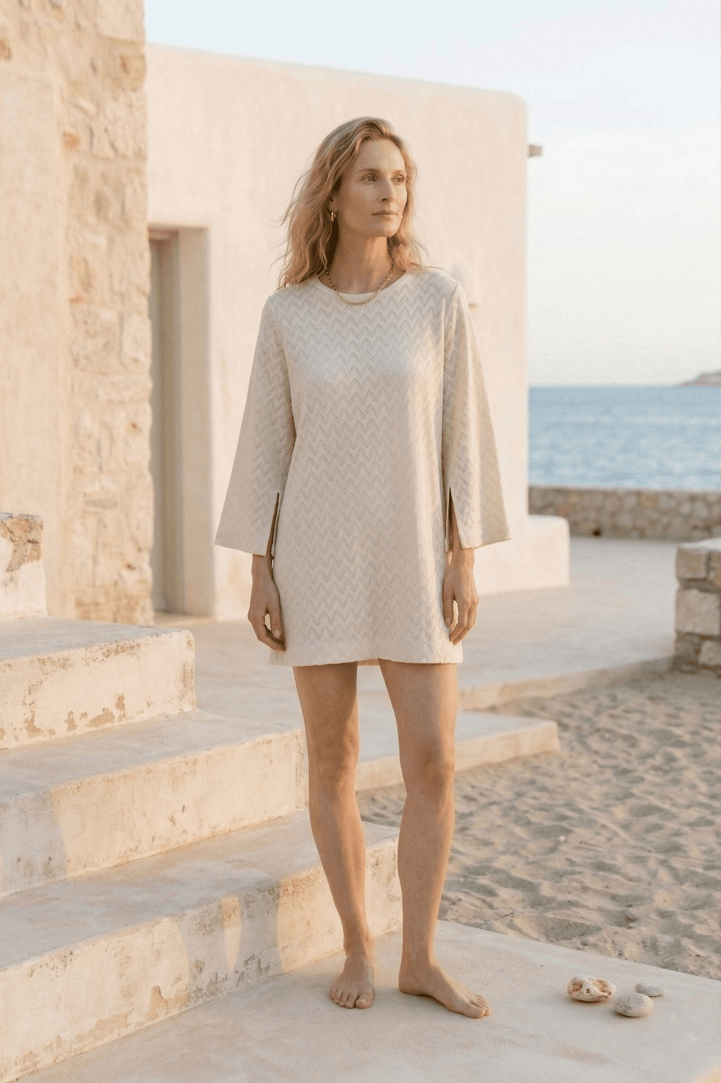 Organic Cotton Crochet Mini Dress worn by a model in a serene coastal setting, featuring flared sleeves in ecru.