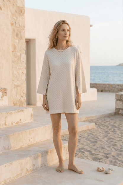 Organic Cotton Crochet Mini Dress worn by a model in a serene coastal setting, featuring flared sleeves in ecru.